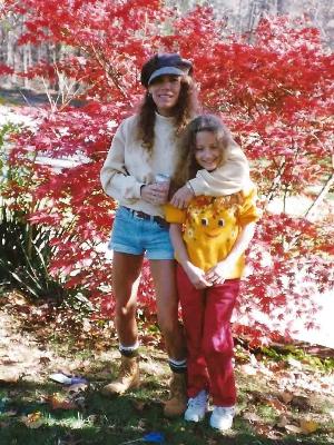 Me with my mom, age 7 or 8. Screen reader caption: A young woman with long curly brown hair, a sweatshirt, and jean shorts standing amidst fall foliage with her arm around a child with long curly hair, red jeans, and an orange sweatshirt with a smiling pumpkin on it.